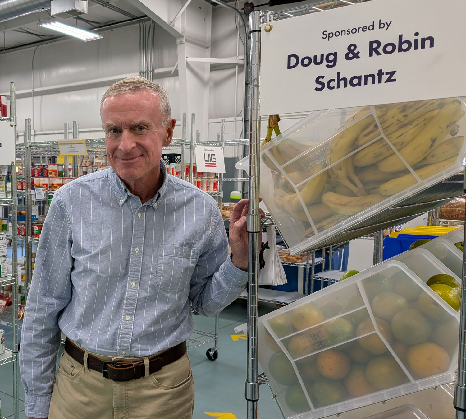 Doug Schantz stands by a shelf of food with a sign that reads, "Sponsored by Doug & Robin Schantz"